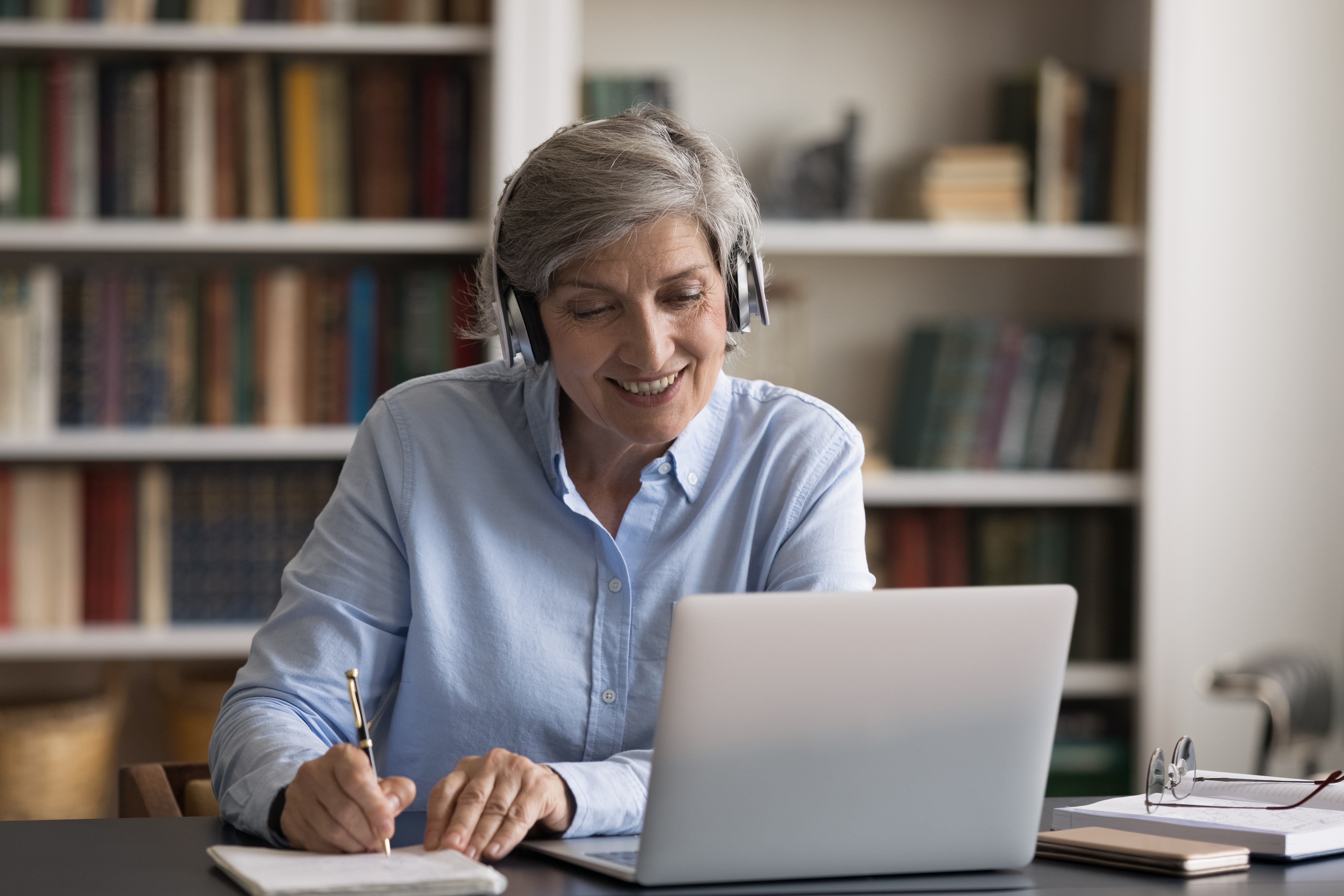 Older woman sitting at computer with headphones taking notes