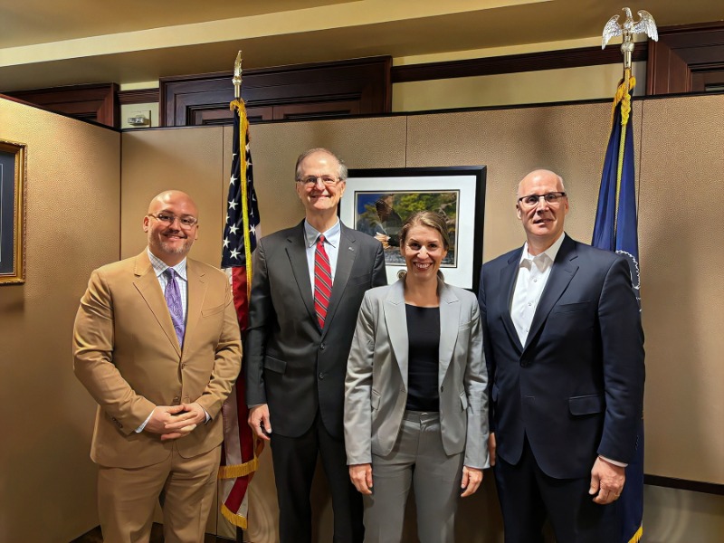Three men and one woman in business suits standing in front of flags
