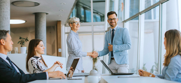 Younger man shaking hands with older woman in front of a board table surrounded by people